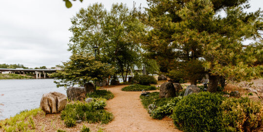 A dirt path goes through a garden of shrubs and trees, decorated with rocks and stone pagodas.